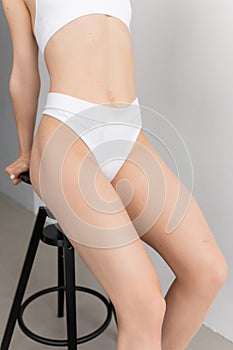 Cropped photo of young woman wearing white seamless underwear bra, thongs, sitting on black stool, holding top of stool.