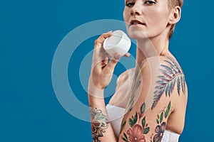 Cropped portrait of beautiful half naked tattooed woman advertising, holding white plastic jar of cream or body lotion