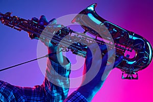 Cropped portrait of jazzman playing saxophone against gradient blue-pink background in neon light. Jazz melodies.