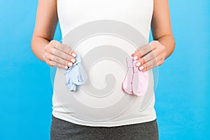 Cropped portrait of pregnant woman in home clothing holding baby pink and blue socks against her belly at blue background. Is it a