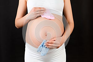 Cropped portrait of pregnant woman in white underwear holding baby pink and blue socks against her belly at black background. Is