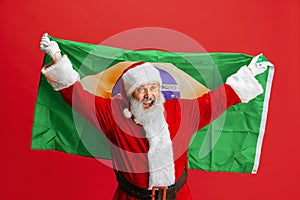 Cropped portrait of senior man wearing Santa Claus costume, holding Brazil flag isolated over red background. Football