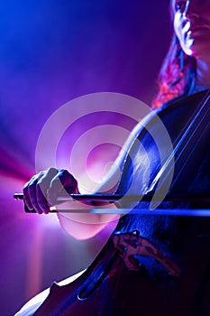 Cropped portrait of talented young woman, musician, cellist performing symphony in purple-blue spotlights on stage with
