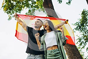 Cropped portrait of two young football fans, man and woman, holding flag of Spain, isolated on park backgroudnd