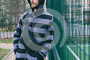 Cropped portrait of young bearded man in striped hoodie with black leather crossbody bag. Street wear, fashion city accessories