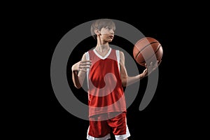 Cropped portrait of young boy, basketball player in red uniform standing, posing with ball isolated over black