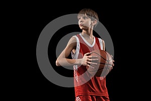 Cropped portrait of young boy, basketball player in red uniform standing, posing with ball isolated over black