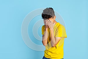 Cropped portrait of young boy in yellow T-shirt covering face with hands isolated over blue background. Sad feelings