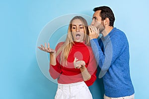 Cropped portrait of young couple, man whispering to shocked woman gossips, news isolated over blue background