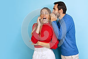 Cropped portrait of young couple, man whispering to woman excited gossips, news isolated over blue background