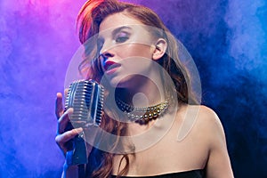 Cropped portrait of young female jazz singer isolated over gradient blue purple background in neon light with stage