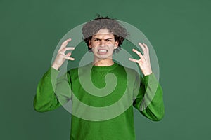 Cropped portrait of young man in green sweater with angry, irritating facial expression isolated over dark green