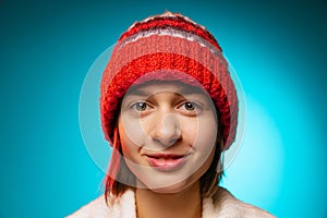 Cropped portrait of young merry girl in red winter hat smiling at camera isolated over blue background