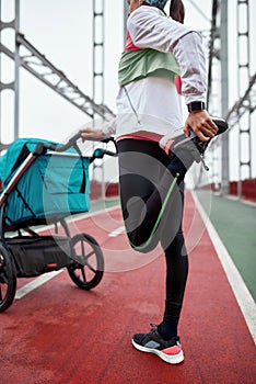 Cropped shot of active mother in sportswear warming up her legs, preparing for a jog on the bridge with a stroller on a