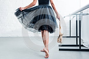 cropped shot of ballerina in black tutu holding pointe shoes in ballet studio