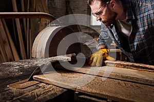 cropped shot of bearded carpenter in protective gloves and googles using machine saw