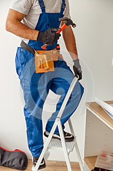 Cropped shot of electrician in uniform using ladder, holding hammer while installing light fitting in new apartment