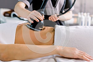 Cropped shot of hands of cosmetologist doing ultrasound cavitation procedure on the belly of woman. Unrecognizable woman