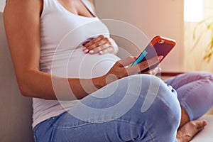 Hands of female touching pregnant belly and using phone at home