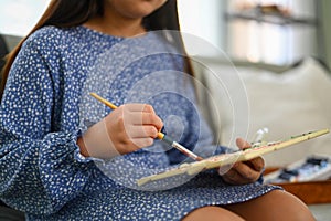 Cropped shot of little girl in casual dress painting picture, enjoying leisure weekend activity at home.