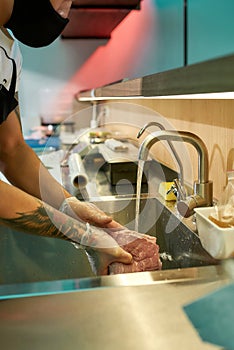 Cropped shot of male cook wearing protective mask and gloves cleaning the raw pork under tap water for cooking