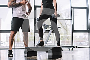 cropped shot of male personal trainer with timer and young sportswoman doing step aerobics exercise with dumbbells at gym