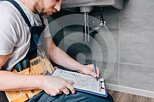 Cropped shot of male plumber with toolbelt writing in clipboard near broken sink