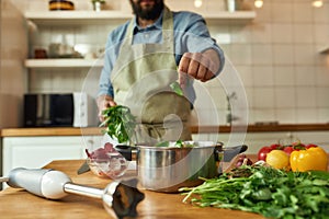 Cropped shot of man, chef cook adding basil leaf to the pot with chopped vegetables while preparing healthy meal, soup