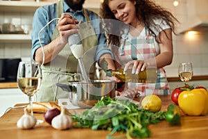 Cropped shot of man, chef cook using hand blender while preparing a meal. Young woman, girlfriend in apron pouring olive
