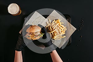 cropped shot of person in gloves holding tasty burger above tray with french fries and glass of beer on black
