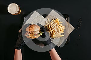 cropped shot of person in gloves holding tasty burger above tray with french fries and glass of beer