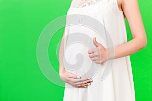 Cropped shot of positive pregnant woman in white dress showing thumb up cool gesture over her baby bump at green background. Happy