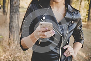 Cropped shot of slender female in black leather jacket holding smartphone at autumn forest background. Beautiful unrecognizable