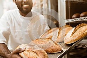 cropped shot of smiling african american baker with tray of fresh loaves of bread