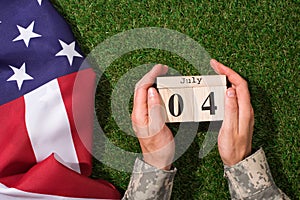 cropped shot of soldier in military uniform holding calendar with 4th july date with american flag on green grass, americas