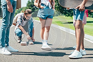 cropped shot of teenagers with books and skateboard waiting for friend tying shoelaces