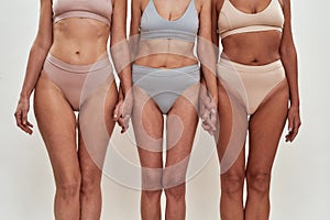 Cropped shot of three middle aged women in underwear holding hands together while standing on light background