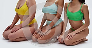 Cropped shot of three young women with different body types in colorful underwear sitting on knees isolated over white