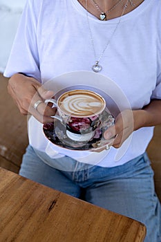 cropped shot of Woman in white shirt holding cup of hot cappuccino on wooden table, top view.