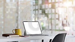 Cropped shot of workspace with blank screen tablet, office supplies and coffee cup on white table with blurred office room