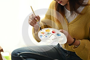 Cropped shot of young female artist is sitting on floor holding tube of oil paint and mixing colors on palette unfinished painting