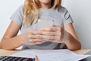 Cropped shot of young female enterpreneur in grey T-shirt, holding smart phone in hands, resting for minute after paper work at of