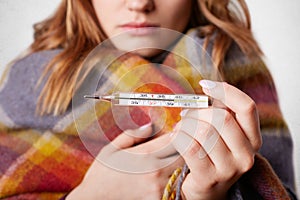 Cropped shot of young female wrapped in warm coverlet, holds termometer which shows high temperature, has flu, over white