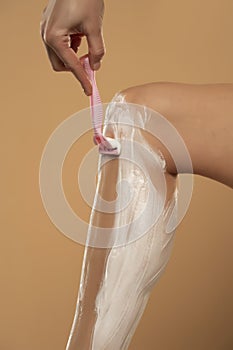 Cropped shot of young woman shaving her leg on beige background