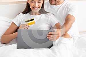 Cropped smiling caucasian young family hugging, sitting in bed and using credit card and laptop for shopping