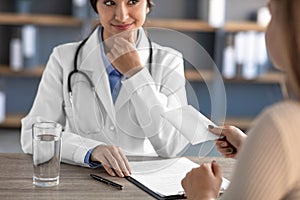 Cropped smiling young indian woman doctor in white coat with stethoscope takes money from patient