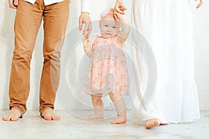 Cropped studio photo of happy, shy, smiling little barefoot baby girl in pink dress and bandelet holding arms together