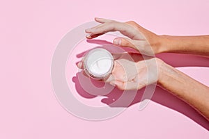 Cropped top view of female hands holding container with cream in hands. Using cosmetics product isolated pastel pink