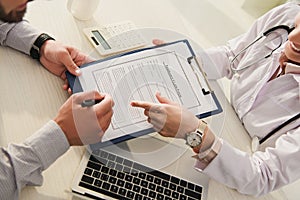 cropped view of client signing insurance claim form while doctor pointing at it in clinic with laptop