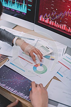 Cropped view of data analysts using charts near calculator and computer monitors